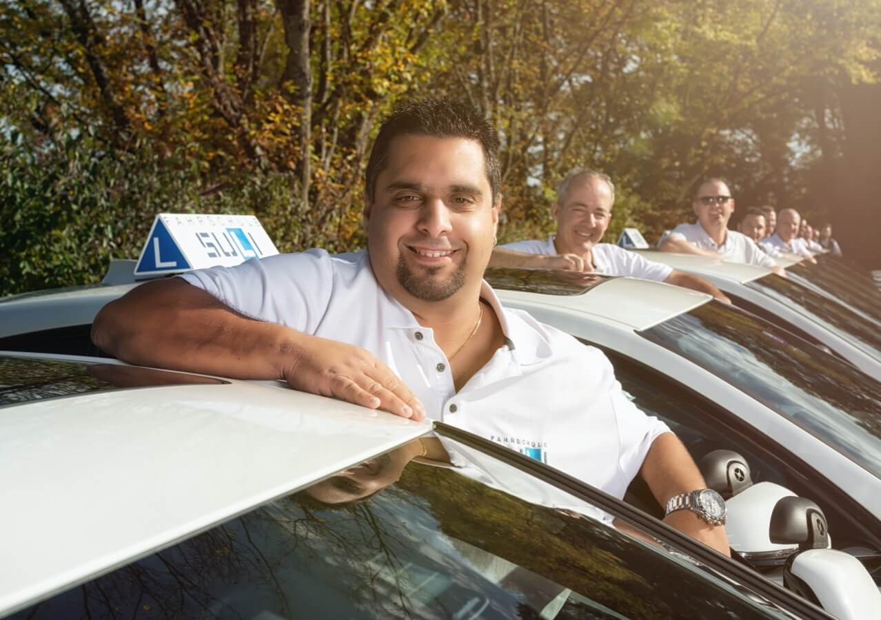 Unsere Fahrlehrer bei ihren Autos, mit Dino im Fokus, der seit dem ersten Tag dabei ist.